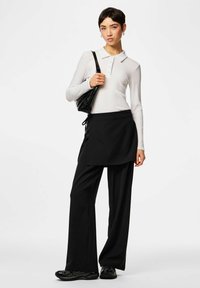 Polo à manches longues blanc, jupe style tablier noire, pantalon large noir, sac noir et baskets noires, mettant en valeur un design de tenue en couches.