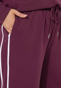 Bordeauxfarbene Jogginghosen aus weichem Stoff mit einem Kordelzugbund und seitlichen Taschen, die mit kontrastierenden weißen Streifen verziert sind.
