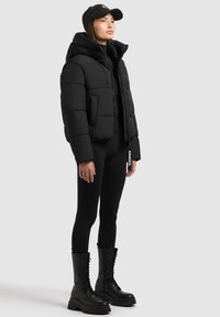 Juoda pūkinė striukė su gobtuvu, užtrauktuko uždarymu ir priekinėmis kišenėmis. Dėvima virš juodų leginsų ir masyvių juodų batų su raišteliais. Ant galvos – juoda kepurė.