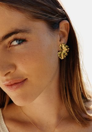 Auskari zelta krāsā ar abstraktu, volāniem līdzīgu dizainu. Auskari ir lieli un izteiksmīgi, ar spīdīgu virsmu, kas atstaro gaismu.