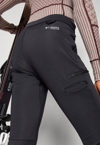 Svarte Columbia Titanium skibukser med en glatt tekstur, sidelommer med glidelås og en tettsittende design, sammen med et mønstrete topp og skiutstyr.