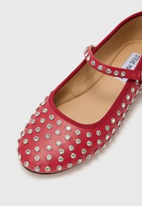 Zapato plano de piel roja con punta redonda, presenta una correa en el empeine y está adornado con pedrería plateada en toda su superficie.