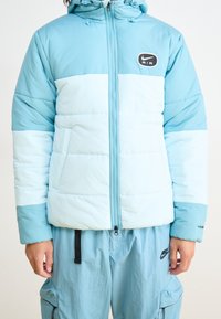 Ljusblått puffjacka med en tvåfärgad design, med dragkedja, huva och Nike Air-logotyp på bröstet. Tillverkad av nylon.