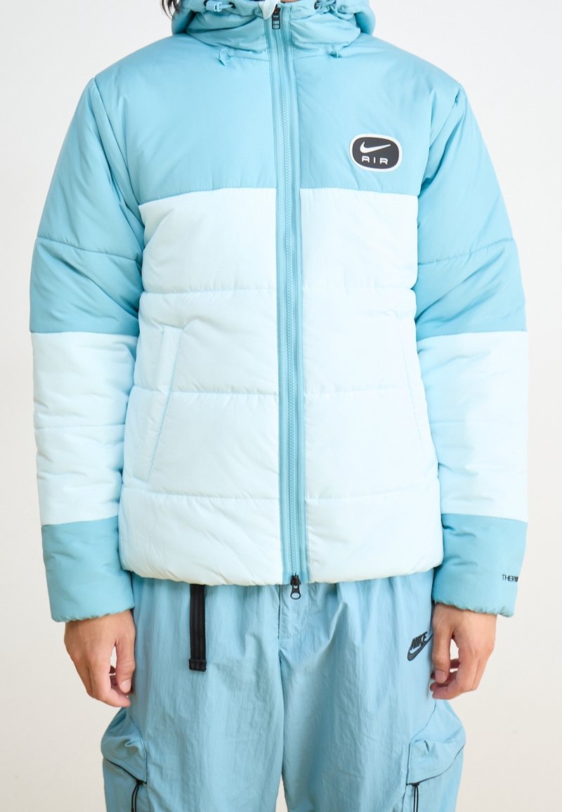 Ljusblått puffjacka med en tvåfärgad design, med dragkedja, huva och Nike Air-logotyp på bröstet. Tillverkad av nylon.