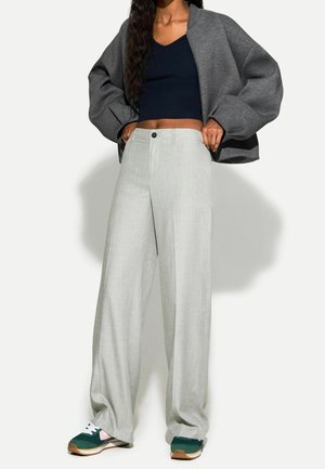 Mujer con pantalones sueltos de color gris claro, top corto azul marino oscuro, chaqueta gris extragrande y zapatillas verdes y blancas, de pie con las manos en los bolsillos.