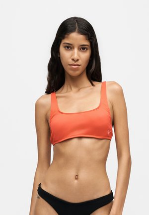 Ung kvinde med langt mørkt hår iført en orange crop top og sorte bikiniunderdele mod en ensfarvet lys baggrund.