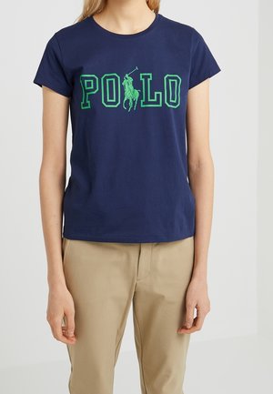 Osoba nosząca granatowy t-shirt z zielonym napisem ""POLO"" i logo konia, w połączeniu z beżowymi spodniami, stojąca na jednolitym tle.