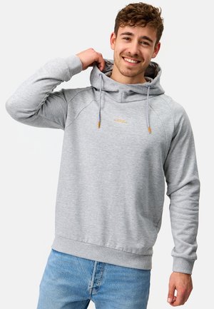 Junger Mann lächelt, trägt ein hellgraues Kapuzensweatshirt mit Kordelzug und blaue Jeans, zieht die Kapuze mit einer Hand zurecht.