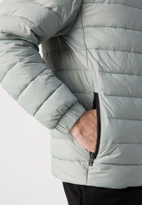 Personne portant une veste matelassée gris clair avec la main dans la poche latérale ouverte, fermeture éclair noire visible et poignet élastique sur la manche.