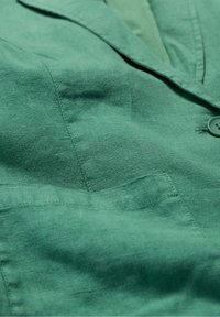 Vihreä pellavablazeri, jossa on teksturoitu kangas, lovettu kaulus, yksi nappi ja rintataskudetaili.