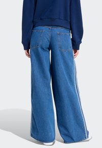 Jeans de mezclilla azul de pierna ancha con dos bolsillos traseros y franjas blancas a los costados. La tela muestra un lavado medio con un acabado ligeramente texturizado.