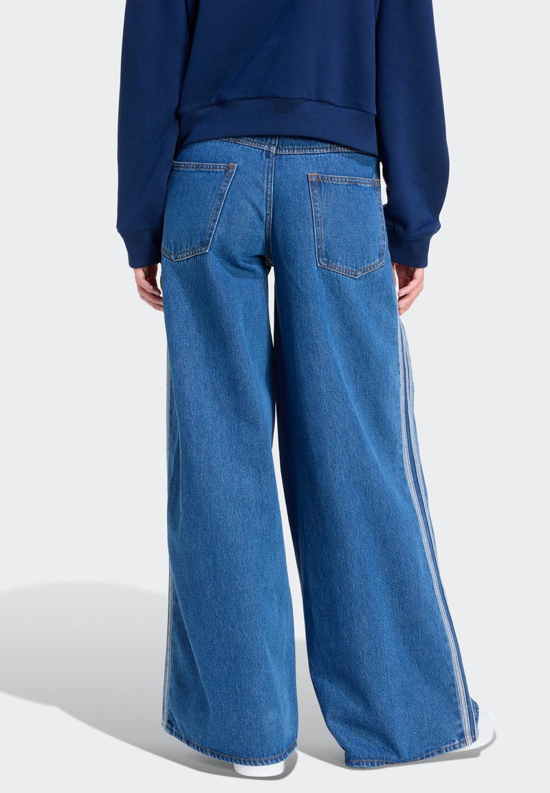 Jean en denim bleu à jambes larges avec deux poches arrière et des bandes blanches sur les côtés. Le tissu présente un lavage moyen avec une finition légèrement texturée.