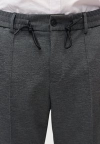 Κοντινό πλάνο σκούρων γκρίζων παντελονιών με υφή, με μαύρο κορδόνι και κούμπωμα με κουμπί, φορεμένα με ανοιχτόχρωμο πουκάμισο που έχει μπηχτεί μέσα.