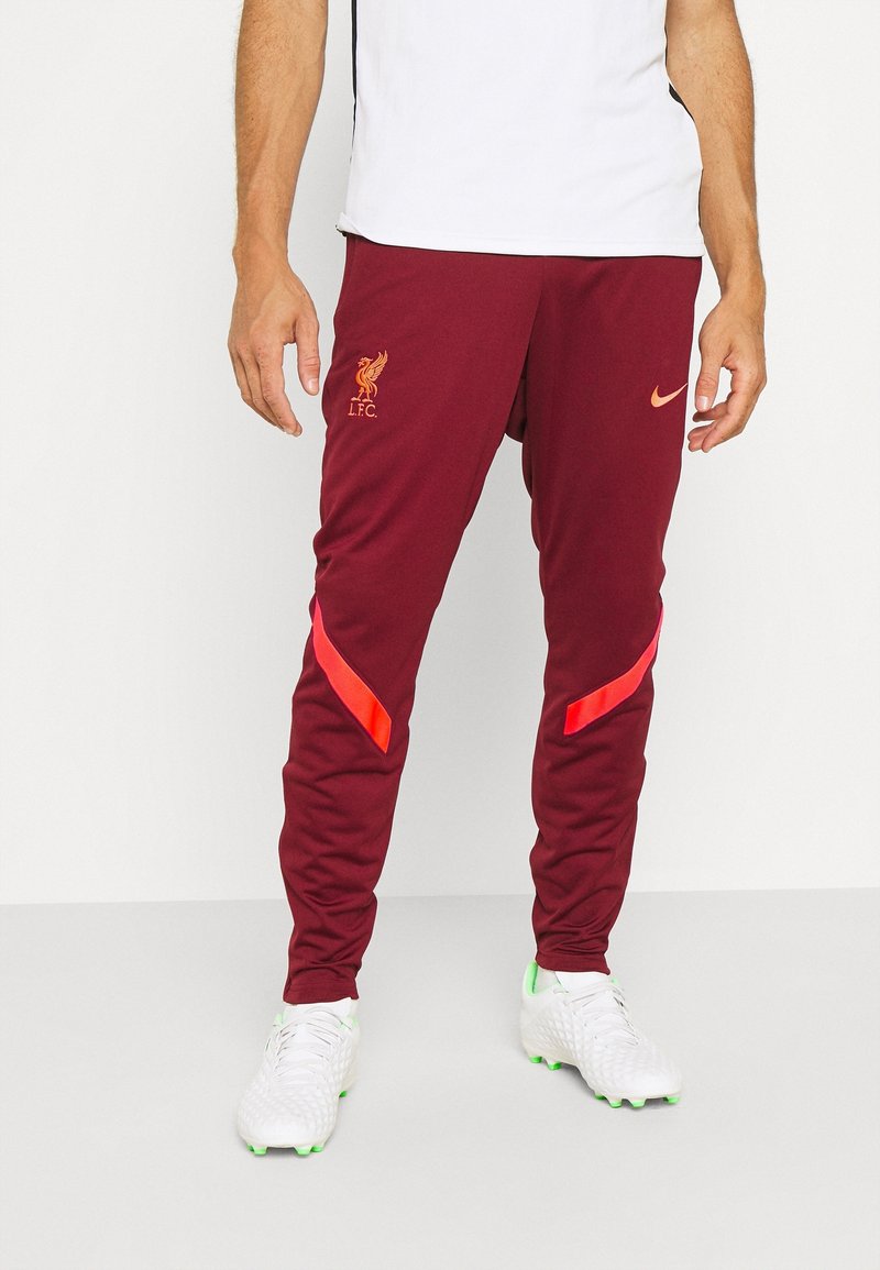 Personne portant un pantalon de sport bordeaux du Liverpool FC avec des bandes rouges et des crampons blancs sur un fond blanc uni.