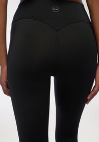 Czarne leginsy z wysokim stanem, z V-kształtnym szwem z tyłu i naszywką z logo na górze. Gładki, elastyczny materiał.
