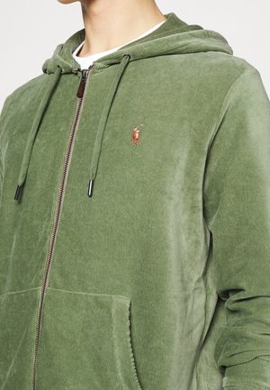 Mężczyzna ubrany w jasnooliwkową bluzę na zamek z sztruksu z małym haftowanym logo i sznurkami przy kapturze.