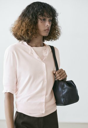 Giovane donna con capelli ricci, che indossa un top rosa chiaro con bottoni e porta una borsa a tracolla in pelle nera.