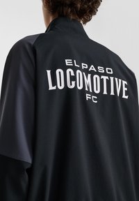 Svart sportjacka med grå paneler, med vit text "EL PASO LOCOMOTIVE FC" som tydligt visas på baksidan. Slät textur.