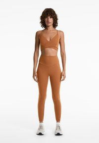 Bruine sport-bh met kruisontwerp en dunne banden, gecombineerd met high-waisted leggings, beide gemaakt van zachte, rekbare stof.