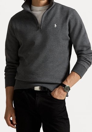 Mężczyzna ubrany w ciemnoszary sweter z zamkiem do połowy, z białym logo Polo, jasnoszarą koszulę pod spodem, czarne spodnie, czarny pasek i czarny zegarek na nadgarstku.