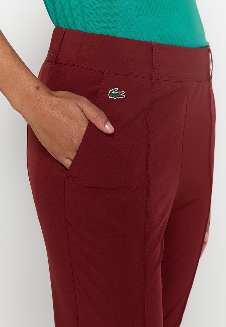 Pantalon bourgogne avec une finition texturée et un logo Lacoste vert. Caractéristiques : poches latérales et une taille ajustée. Matériau : tissu extensible.