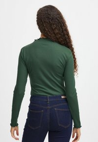 Grön ribbad långärmad topp med volangkrage, parat med mörkblå jeans med kontrasterande sömmar och bakfickor.