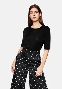 Schwarzes Kurzarmoberteil mit rundem Halsausschnitt, kombiniert mit hochgeschnittenen schwarzen weiten Hosen mit großen weißen Pünktchen.