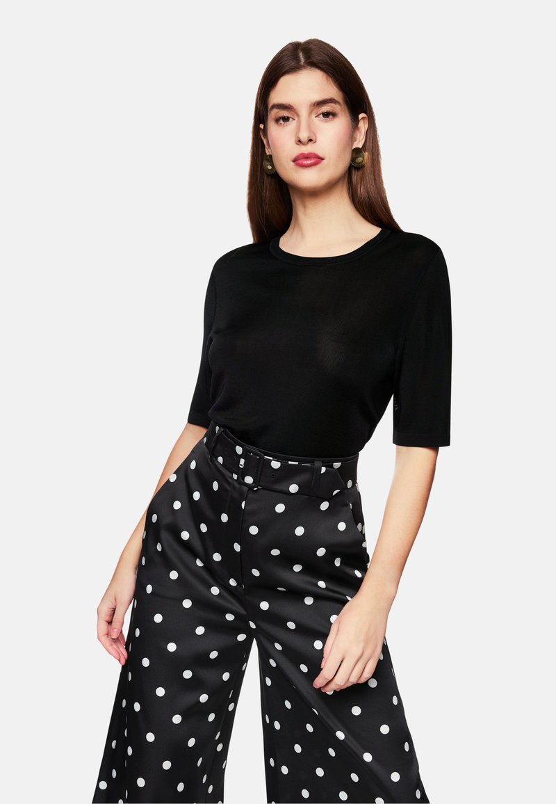 Schwarzes Kurzarmoberteil mit rundem Halsausschnitt, kombiniert mit hochgeschnittenen schwarzen weiten Hosen mit großen weißen Pünktchen.