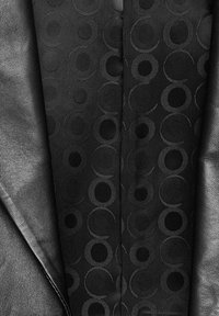 Zwarte tuxedo jasje met een gladde leren afwerking, gecombineerd met een gestreepte zwarte vest dat cirkelvormige ontwerpen in verschillende texturen weergeeft.
