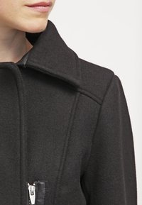 Nainen, jolla on päällään musta villakangastakki, jossa on leveä kaulus ja näkyvä hopeinen vetoketjullinen tasku rinnassa, valkoista taustaa vasten.