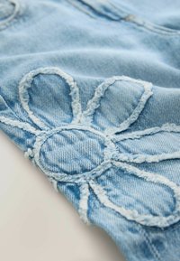 Šviesiai mėlynas džinsinis audinys su siuvinėta gėlės išdėstymu, kuriame matomi ryškūs žiedlapiai ir apskritas centras. Kraštai atrodo nudrengti.