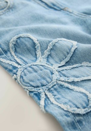 Ljusblå denimtyg med en sydd blomdesign med framträdande kronblad och en cirkulär mitt. Sliten textur längs kanterna.