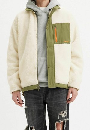 Homme portant une veste sherpa crème avec des bordures vert olive et une poche poitrine par-dessus un sweat à capuche gris, associée à un jean noir déchiré.