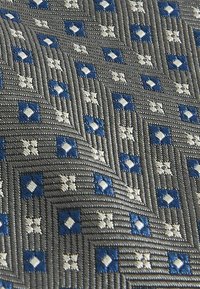 Geometrikus mintázatú textil, amely kék négyzetekből és fehér rombuszokból áll, textúrált szürke háttérrel és bordázott felülettel.