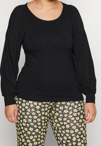 Maglietta nera a maniche lunghe con spalle arricciate, abbinata a leggings aderenti con un motivo floreale giallo su sfondo nero.