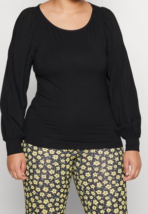 Schwarzes Langarmoberteil mit gerafften Schultern, kombiniert mit enganliegenden Leggings, die ein gelbes Blumenmuster auf schwarzem Hintergrund zeigen.