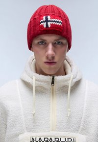 Gorro de punto rojo con un patrón nórdico y una cruz blanca y azul marino. Combinado con una chaqueta de forro polar crema con cremallera frontal y parche de logo.