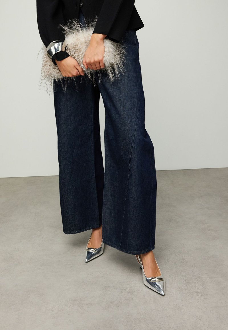Jean denim foncé à jambes larges associé à des escarpins argentés à bouts pointus. Une pochette argentée duveteuse tenue dans une main, haut noir avec des accents de poignets visibles.