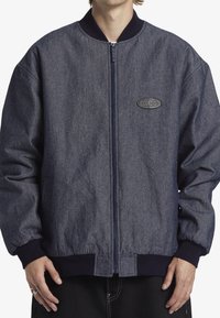 Denim bomberjacka i mörkblått, med dragkedjestängning, ribbade cuffs och nederkant, samt en strukturerad logotyp patch på bröstet.