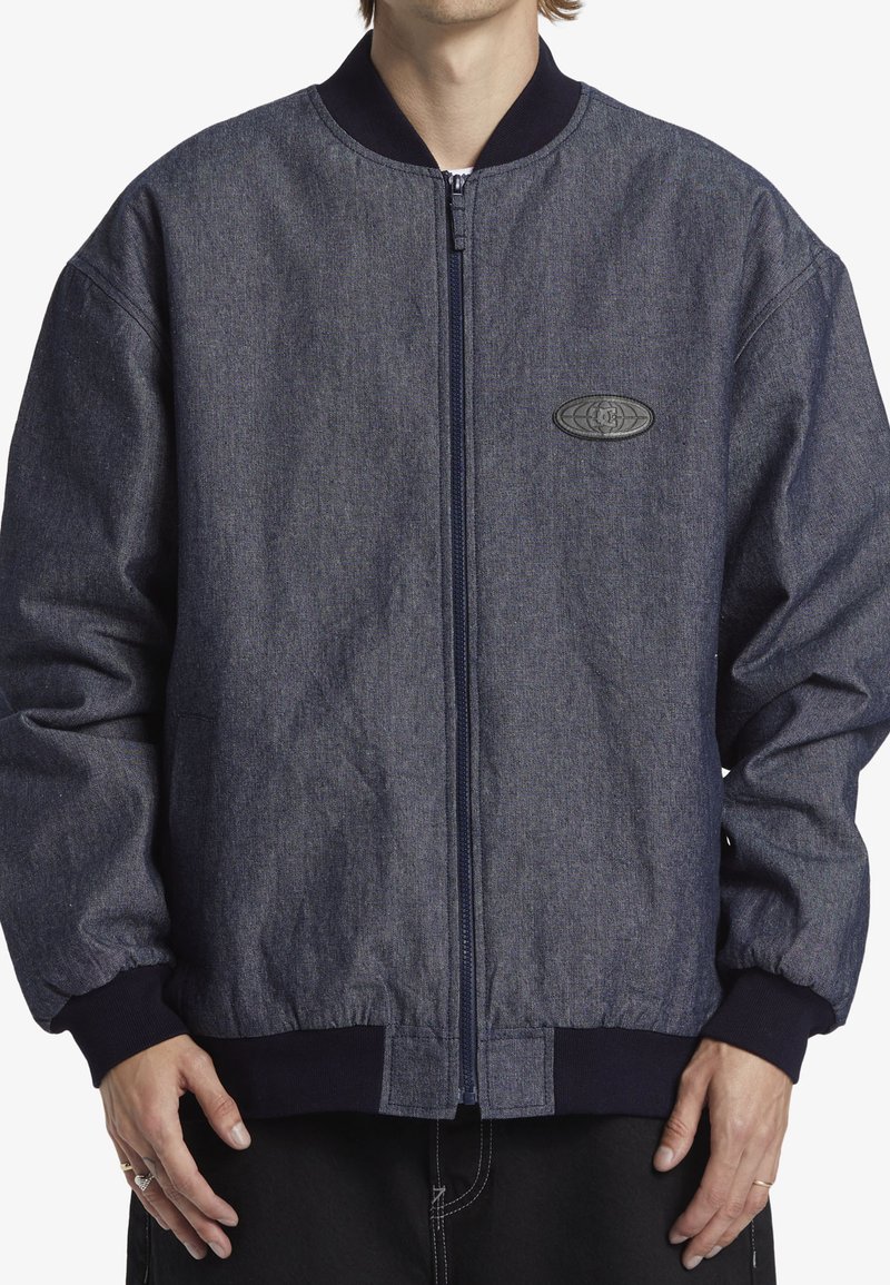Denim bomberjacka i mörkblått, med dragkedjestängning, ribbade cuffs och nederkant, samt en strukturerad logotyp patch på bröstet.