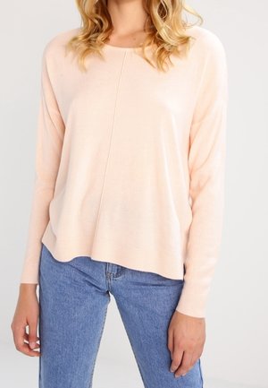 Žena v světlém růžovém svetru s dlouhým rukávem a modrých džínách stojí před jednobarevným světlejším pozadím.