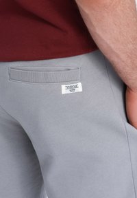 Pantalones de chándal grises hechos de una tela suave, con un bolsillo trasero con ribete y una etiqueta blanca con texto. Combinados con una parte superior color burdeos.