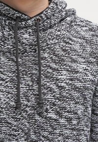 Grau-weiß gemusterter Hoodie mit Krempenkragen, ausgestattet mit dunkelgrauen Kordeln und strukturiertem Stoff, der eine gemischte, gestrickte Optik hervorhebt.