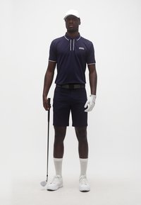 Άνδρας με ναυτικό γκολφ σύνολο, λευκό καπέλο, γάντι, κάλτσες και παπούτσια, κρατώντας ένα γκολφ ρόπαλο, στέκεται μπροστά σε απλό λευκό φόντο.