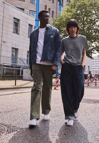 Veste en jean, t-shirt blanc, pantalon vert olive et baskets blanches sur une personne ; t-shirt rayé, jean large bleu et baskets blanches sur une autre.