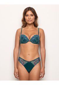 Donna che indossa un reggiseno turchese e un perizoma abbinato con ricami floreali rosa, in piedi contro uno sfondo chiaro e semplice, frontale.