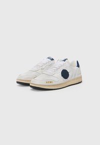 Hvite sneakers laget av lær og tekstil, med marineblå detaljer, rund logo-patch, og en teksturert gummisåle med gul merking.