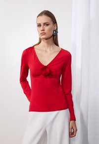 Rote Langarmbluse mit tiefem V-Ausschnitt und floralen Designdetails in der Mitte. Glatte Textur; kombiniert mit weißen Hosen.