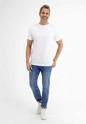 Weißes Baumwoll-T-Shirt mit kurzen Ärmeln, kombiniert mit hellblauen Jeans. Das Model trägt weiße Sneaker und steht vor einem neutralen Hintergrund.