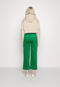 Haut à capuche beige clair avec une poche au dos, associé à un pantalon large en velours côtelé vert. Les chaussures sont des baskets gris clair.